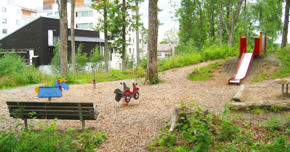 Naturlekplats med barkflis där det finns en fjädergunga, gungdjur och en röd rutschkana på en kulle. Träd och buskar omger lekplatsen och en parkbänk står vid sidan. I bakgrunden syns flervåningshus.