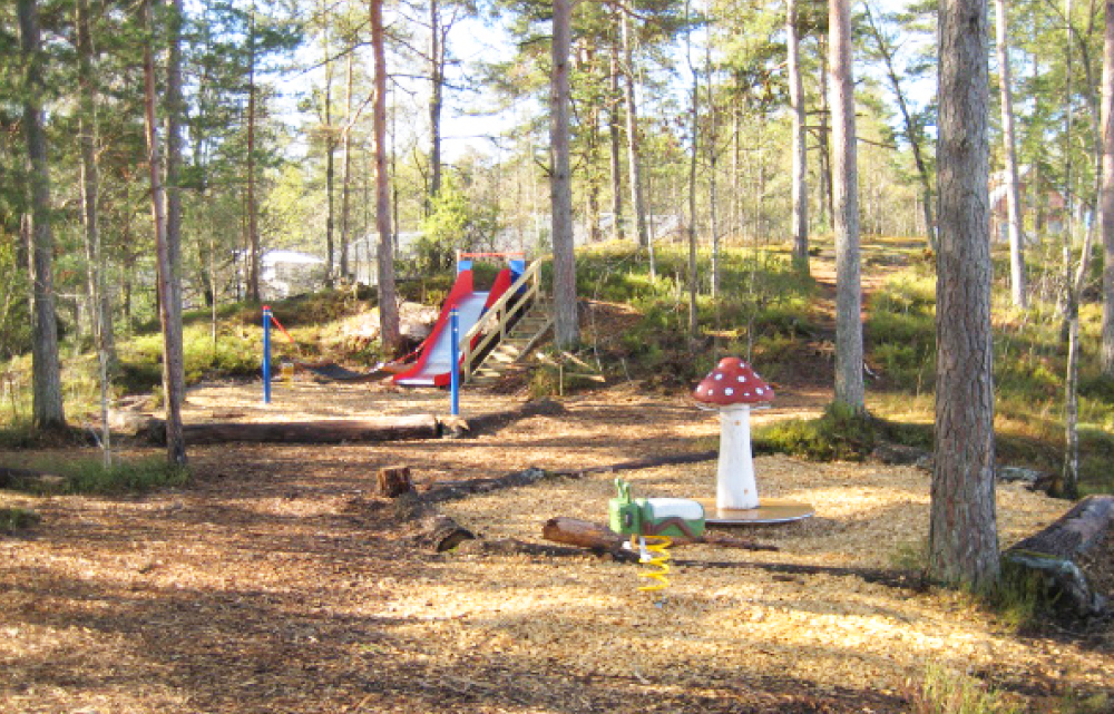 Lekplats mitt i naturen. Omringad av träd finns en rutschkana. I förgrunden syns en svamp och en fjädergunga som liknar en insekt.