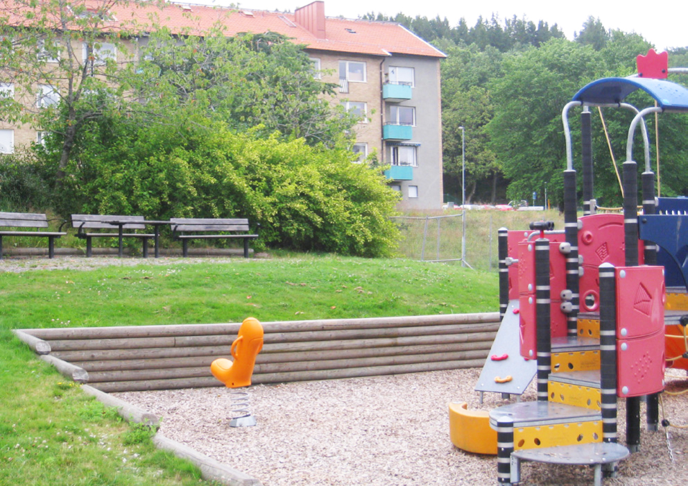 Bostadshus syns i bakgrunden. En del av en lekplatsmodul med trappor till höger. En fjädergunga finns på plats.