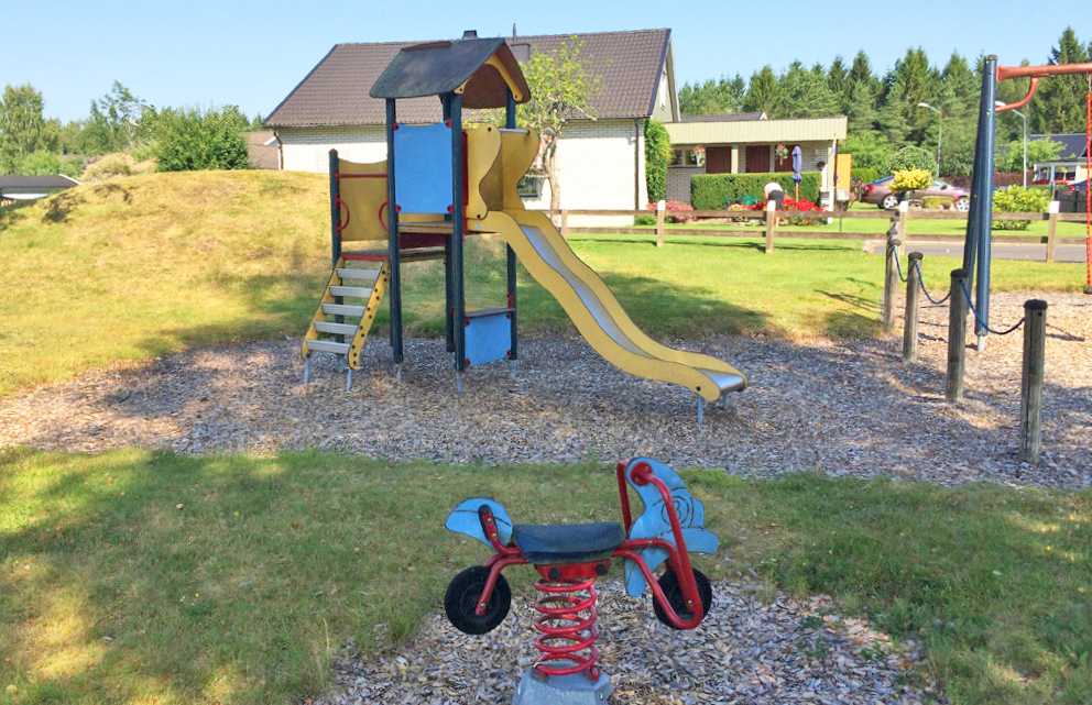 Lekplats med ett blått och gult klättertorn med rutschkana samt en röd gungcykel på fjäder i förgrunden. I bakgrunden syns villor och gröna gräsytor.