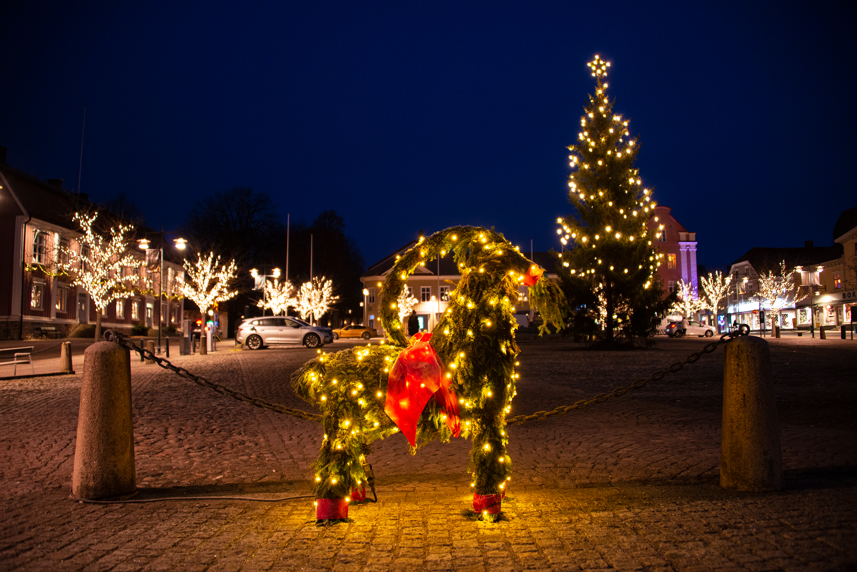 Julbock på Stora Torget i Alingsås i kvällstid. Bakom syns en stor julgran med stjärna i toppen samt träd med belysning.