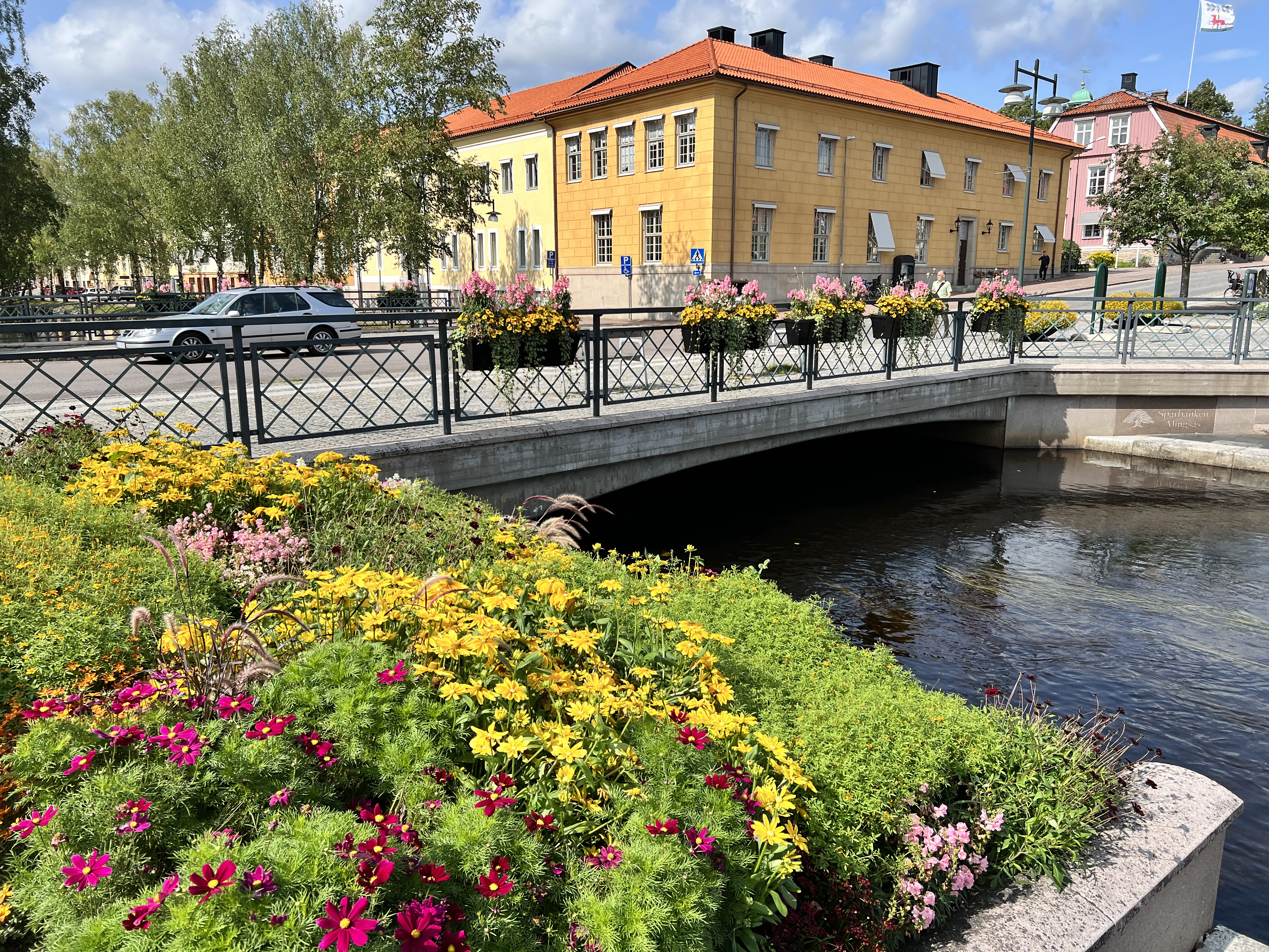 Bro vid Lillån i Alingsås med säsongsplantering i förgrunden. Ett gult, putsat stenhus i bakgrunden.